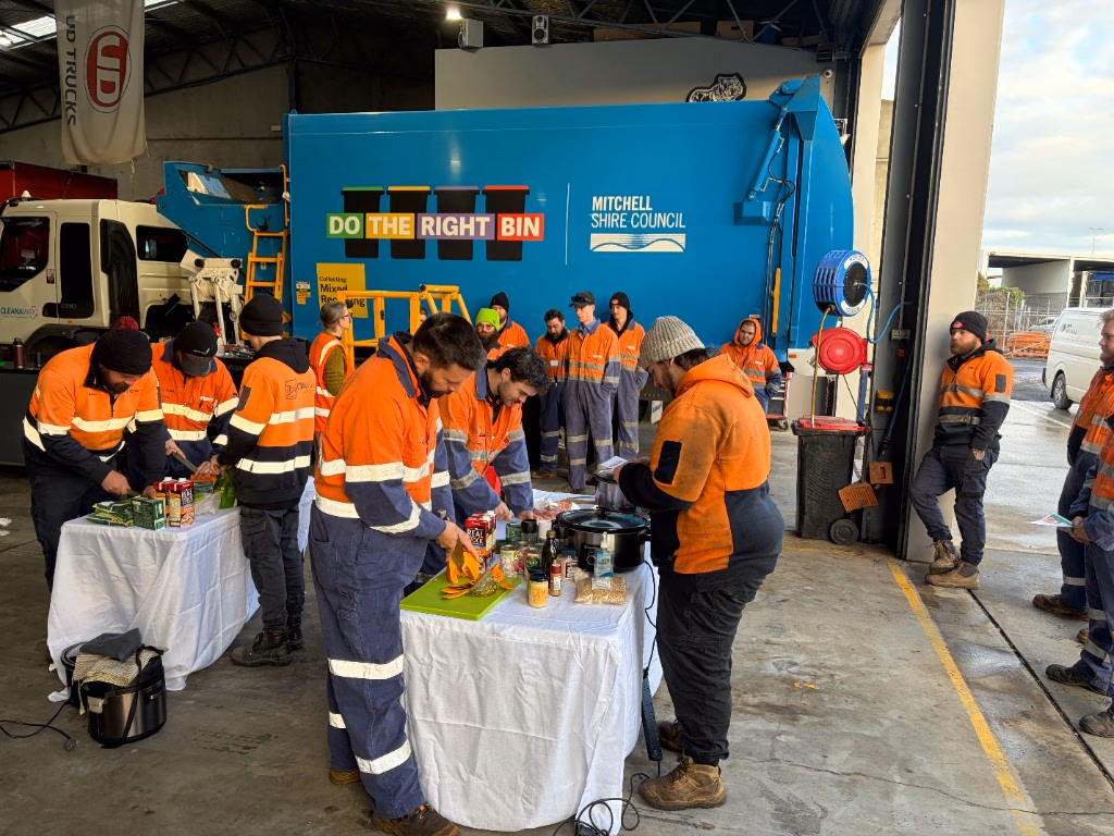 Workers in orange high-visibility clothing chopping and cooking at cloth-covered tables in front of a blue Mitchell Shire Council garbage truck.