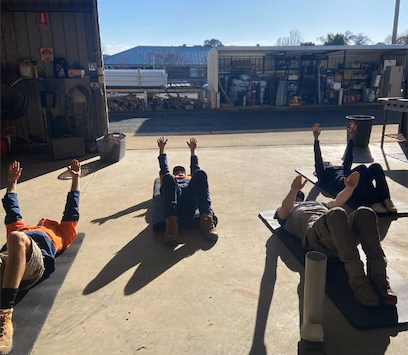 Four workers lying on their backs on mats outside a shed, stretching with their legs raised.