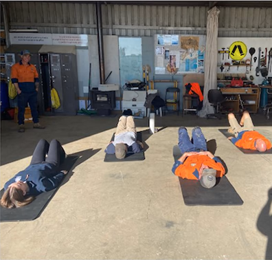 People lying on black mats inside a workshop while a person in orange high-vis stands near the doorway.