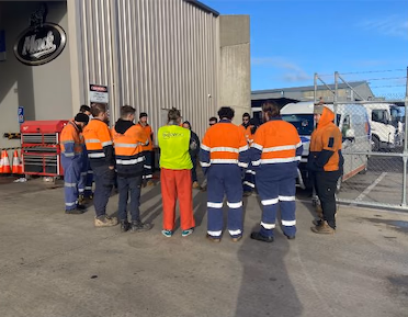 Workers in orange high-visibility clothing standing in a circle outside a workshop.