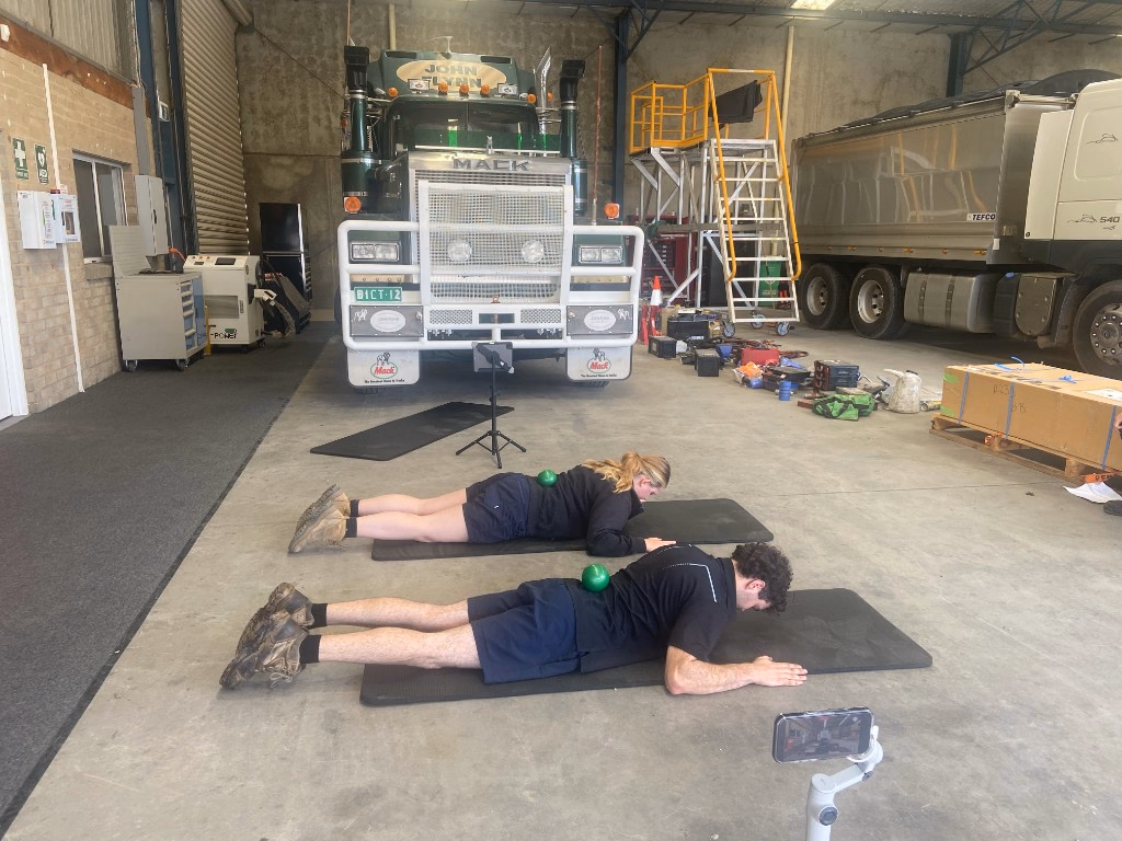 Two people lying face down on black exercise mats in a truck workshop, each with a green ball on their back, and a phone recording from a small tripod.